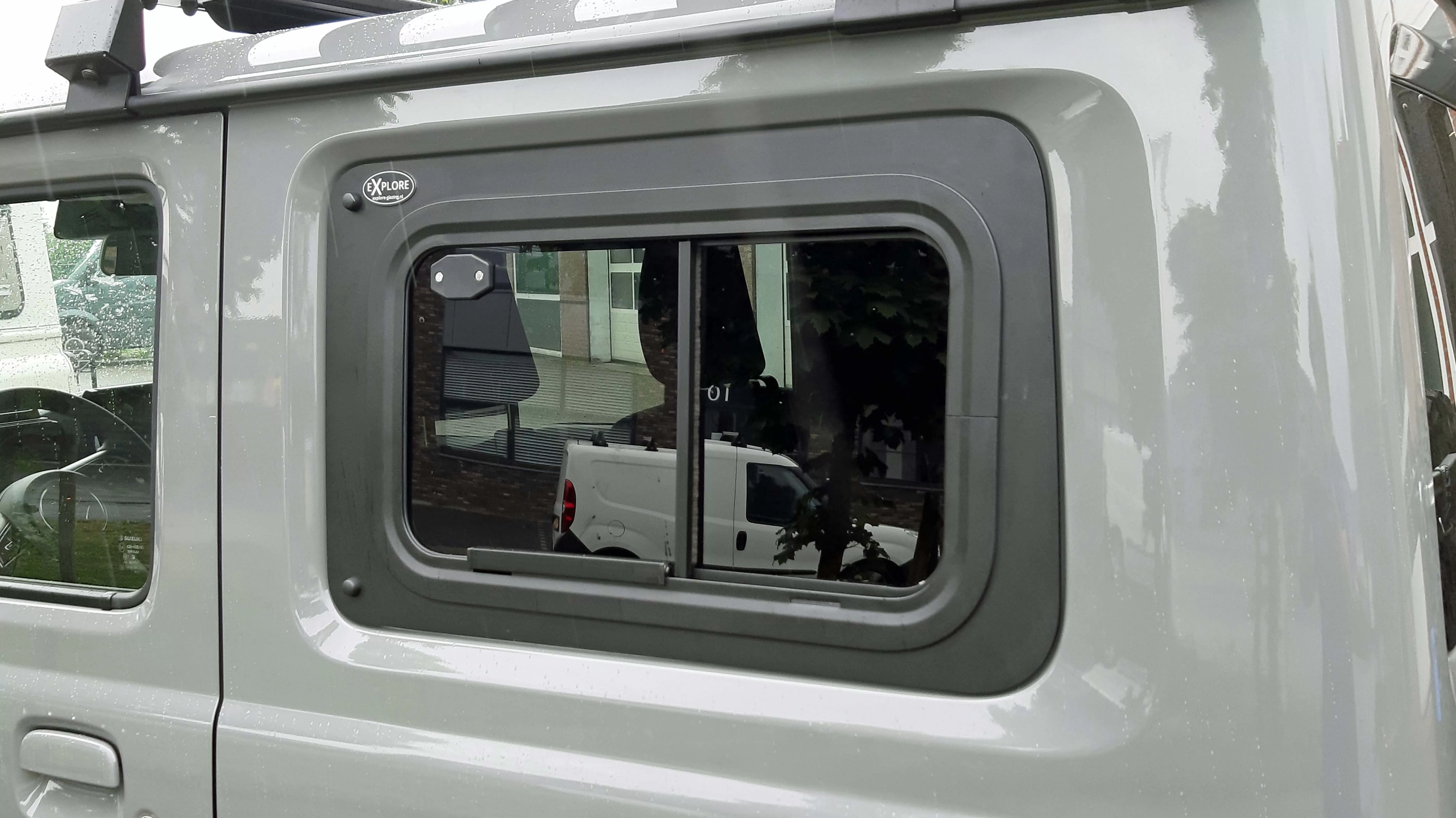 Close-up view of Explore Glazing's exterior sliding window fitted on a Suzuki Jimny GJ (JB74), HJ, or GLX 3-door, featuring a sleek design and rain droplets on the surface, reflecting the urban environment.